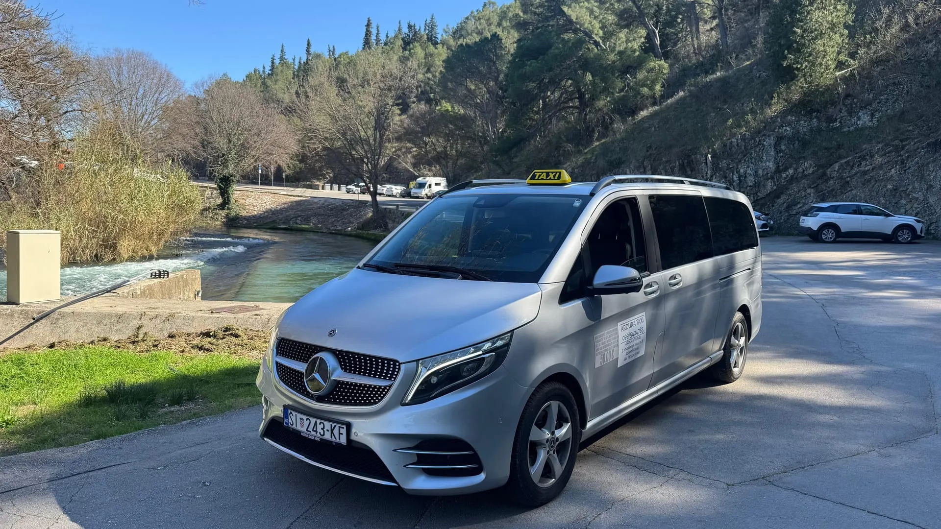 Silver Mercedes V-Class taxi van with a TAXI roof sign parked by the Krka river